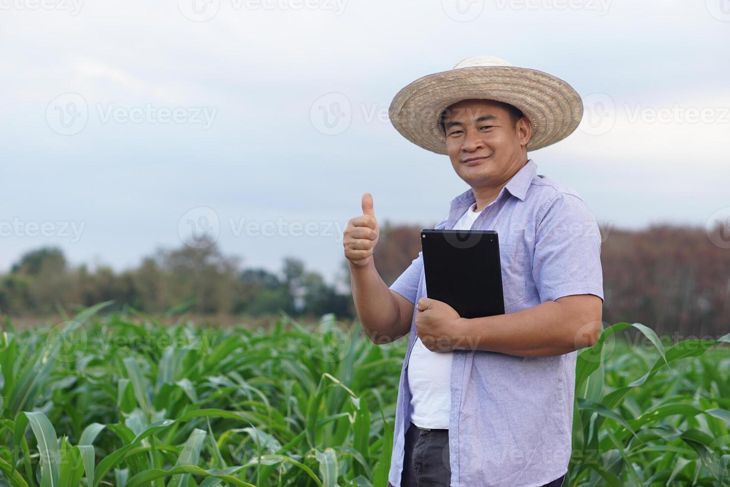 Handsome Asian man farmer is at garden, wears hat, holds smart tablet, thumbs up to show his satisfaction in crops. Look at camera. Feel confident, proud in crops. Concept , Agriculture job. photo