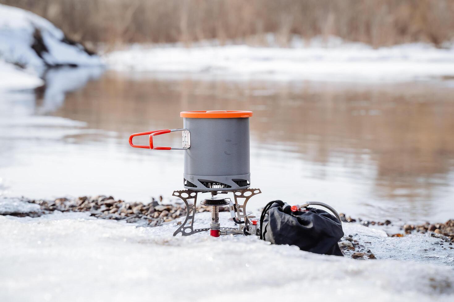 concepto de turista platos, un maceta con un tapa soportes en un gas quemador en el banco de el río, hirviendo agua en un caminata en un cacerola. invierno caminata en el montañas. foto