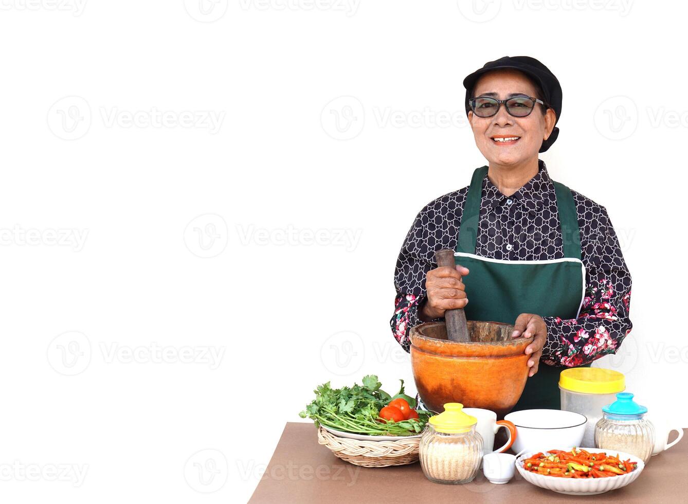 Happy Asian senior woman is cooking, wear chef cap and apron, holds pestle, mortar and plate of chillies, isolated on white background. Concept, Cooking for family. Thai elderly kitchen lifestyle. photo