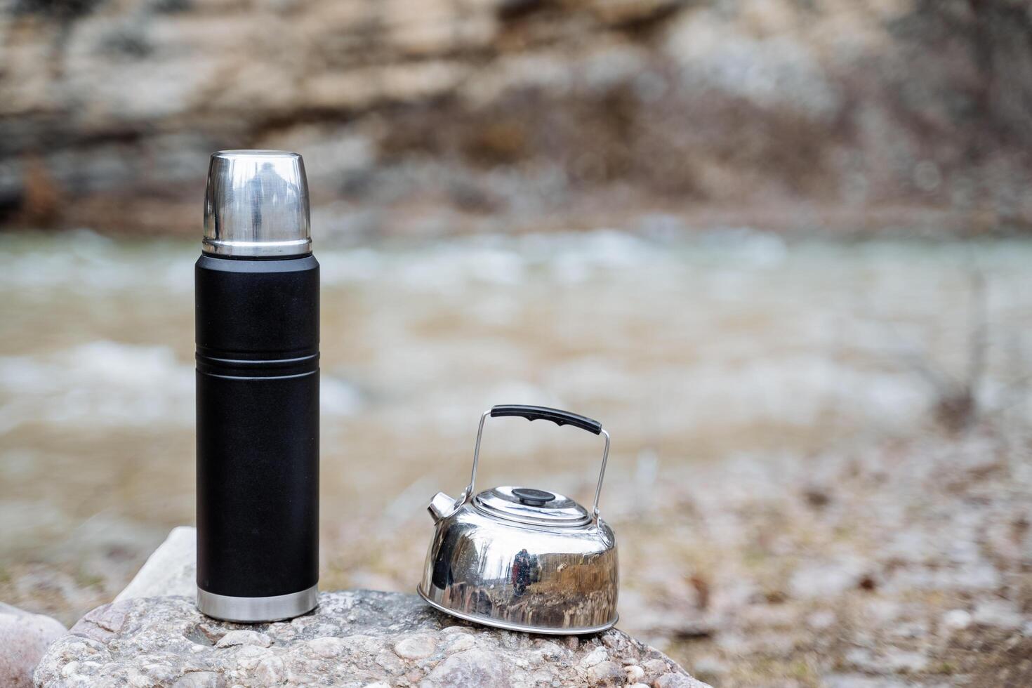 Tourist dishes stand on the stones against the background of the river, a metal kettle glitters, containers for hot drinks. photo