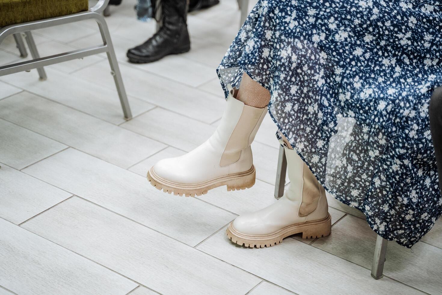 un niña en un azul vestir con un modelo de flores se sienta en un silla en blanco botas. elegante De las mujeres Zapatos con grueso suelas el muchachas pies son vestido en zapatos. foto