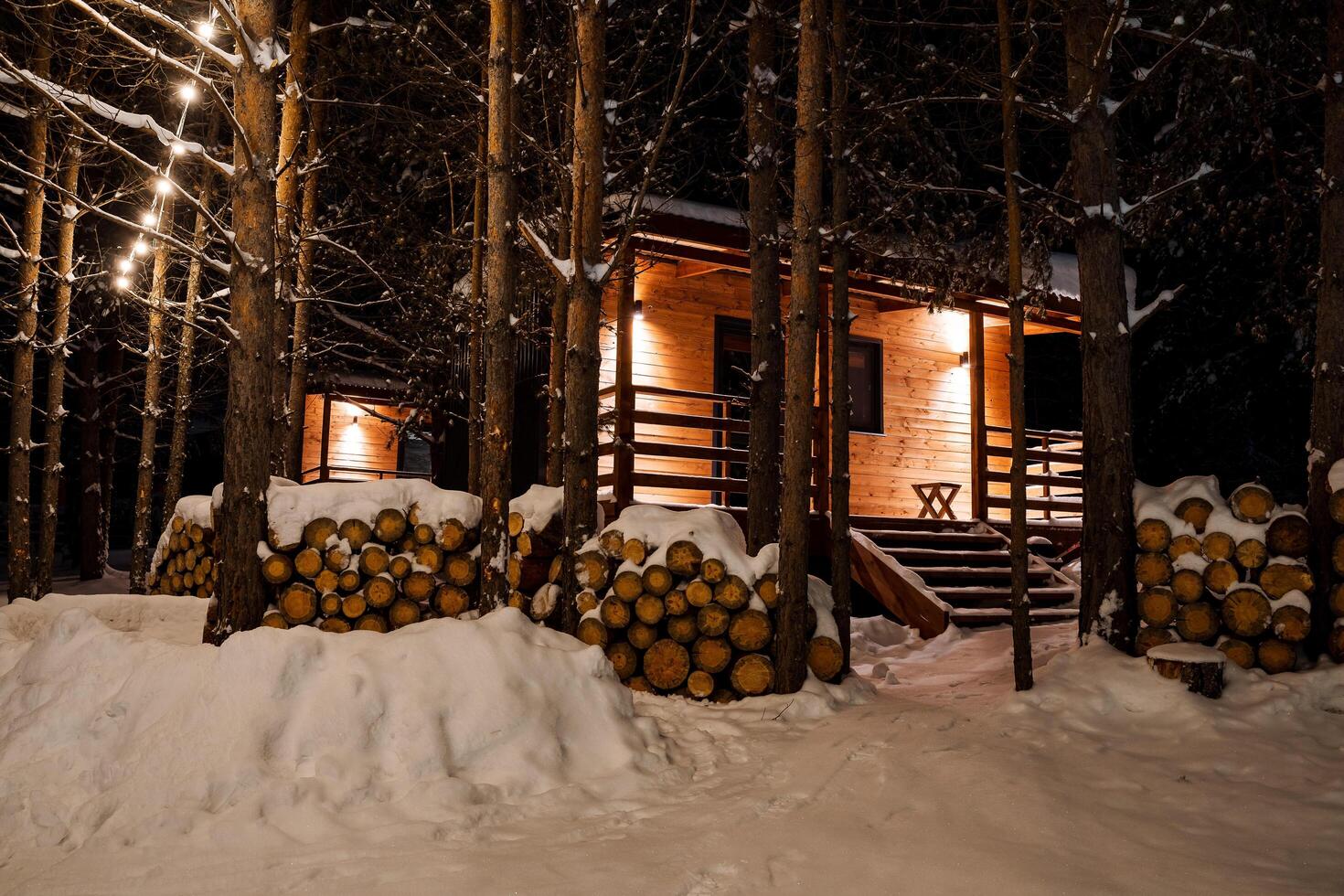 A mesmerizing view of a garland hung in the trees in front of the house. New Year's mood and decorations. Modular country house in the middle of the forest. Chopped wood for fireplace and sauna photo