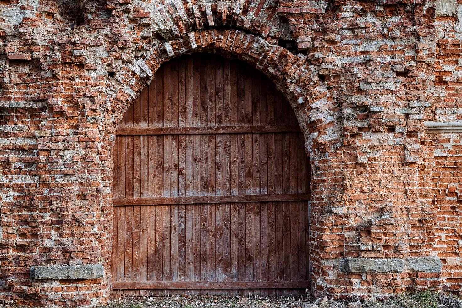 A powerful wooden door is closed, an arched passage in the wall of an old building, an ancient castle is built of red brick, a ruined structure photo