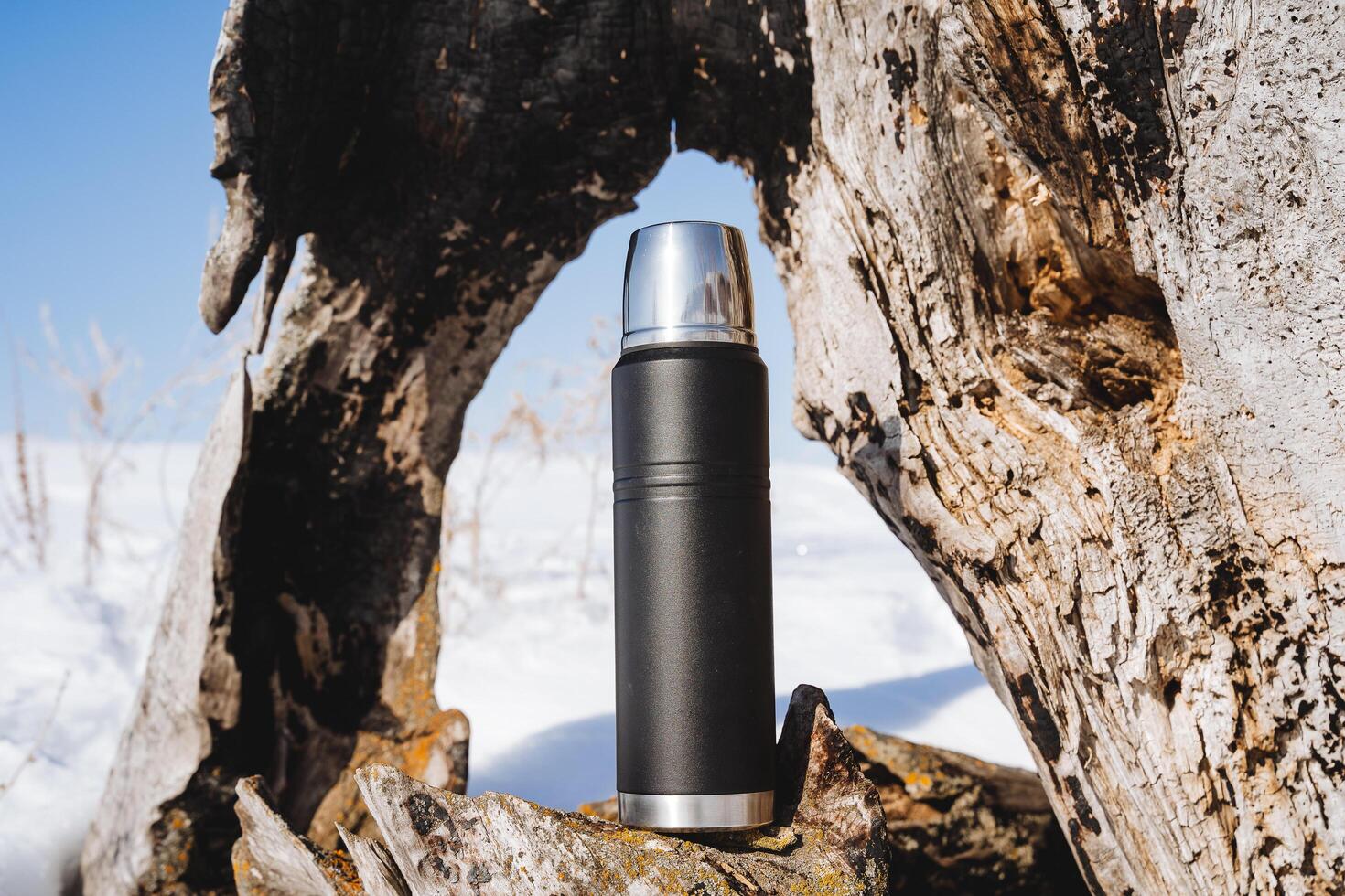 Thermo bottle for hot drinks, thermos stands on a wooden stump, hot tea is poured into a thermos, warm up in winter with a hot drink in nature. photo