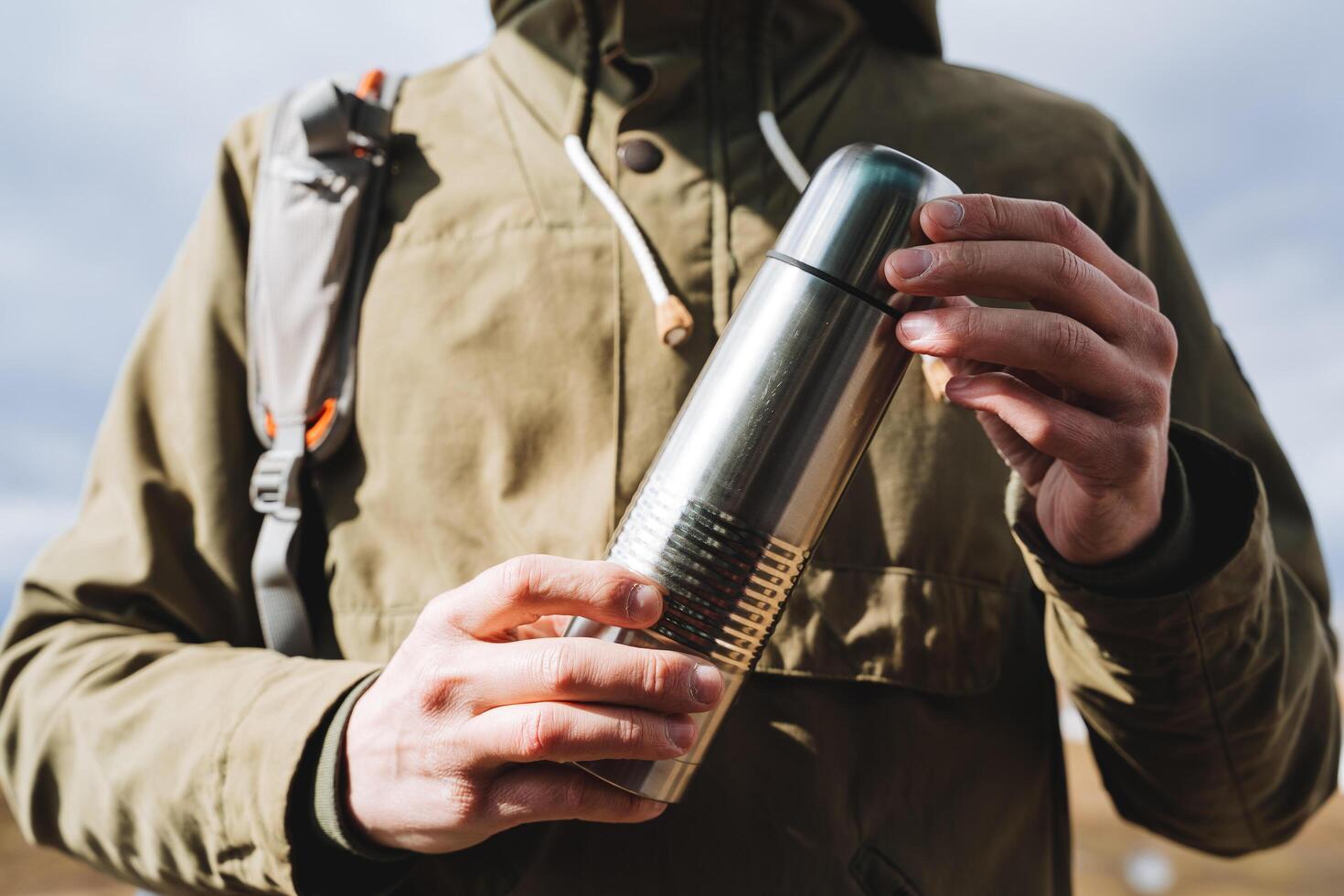 Vacuum silver thermos is held in the hands of a man, the concept of a camping kitchen, a metal bottle for hot drinks, equipment for a hike. photo