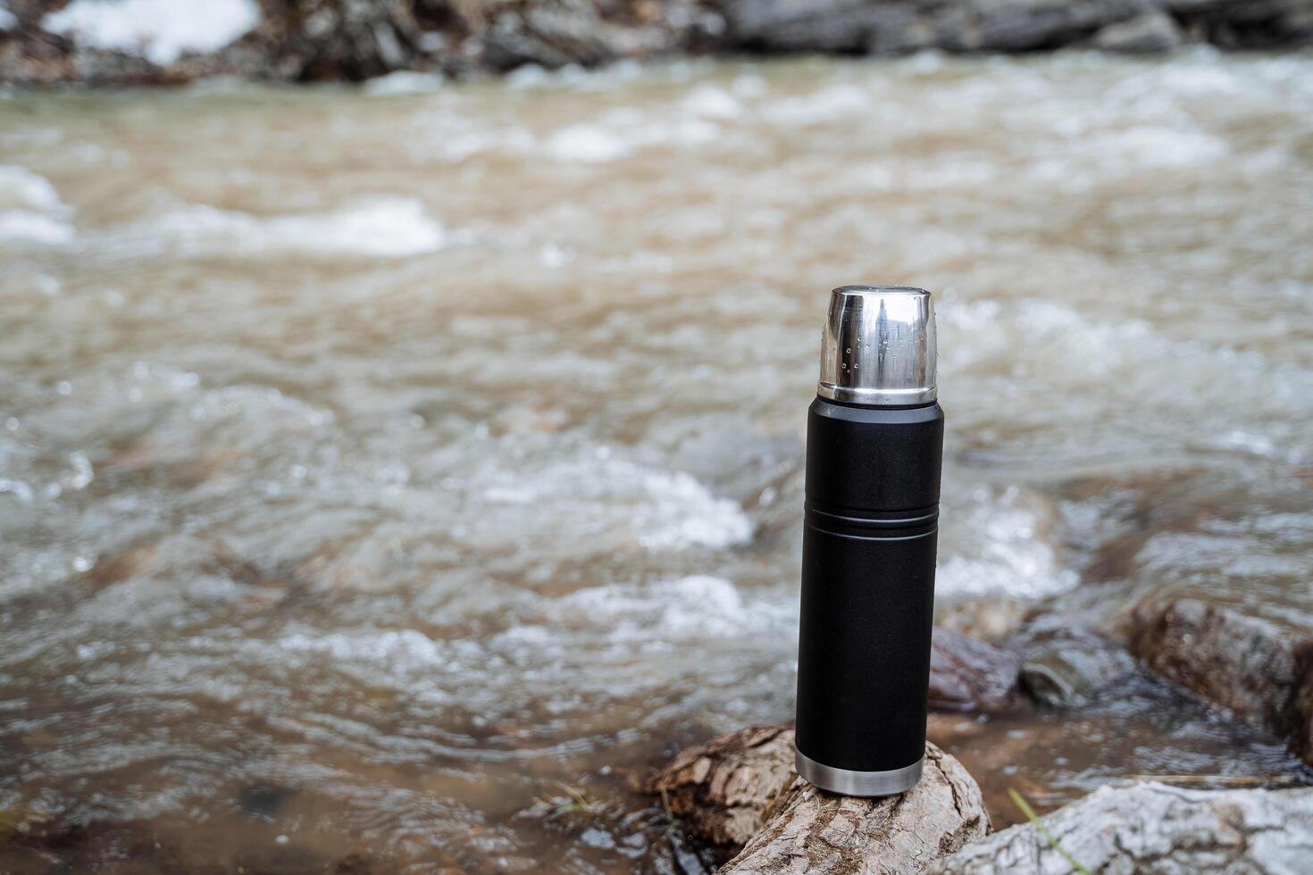 Vacuum thermos stands on a stone against the background of the river, tourist dishes, Spring trekking in nature. Black metal thermos. photo