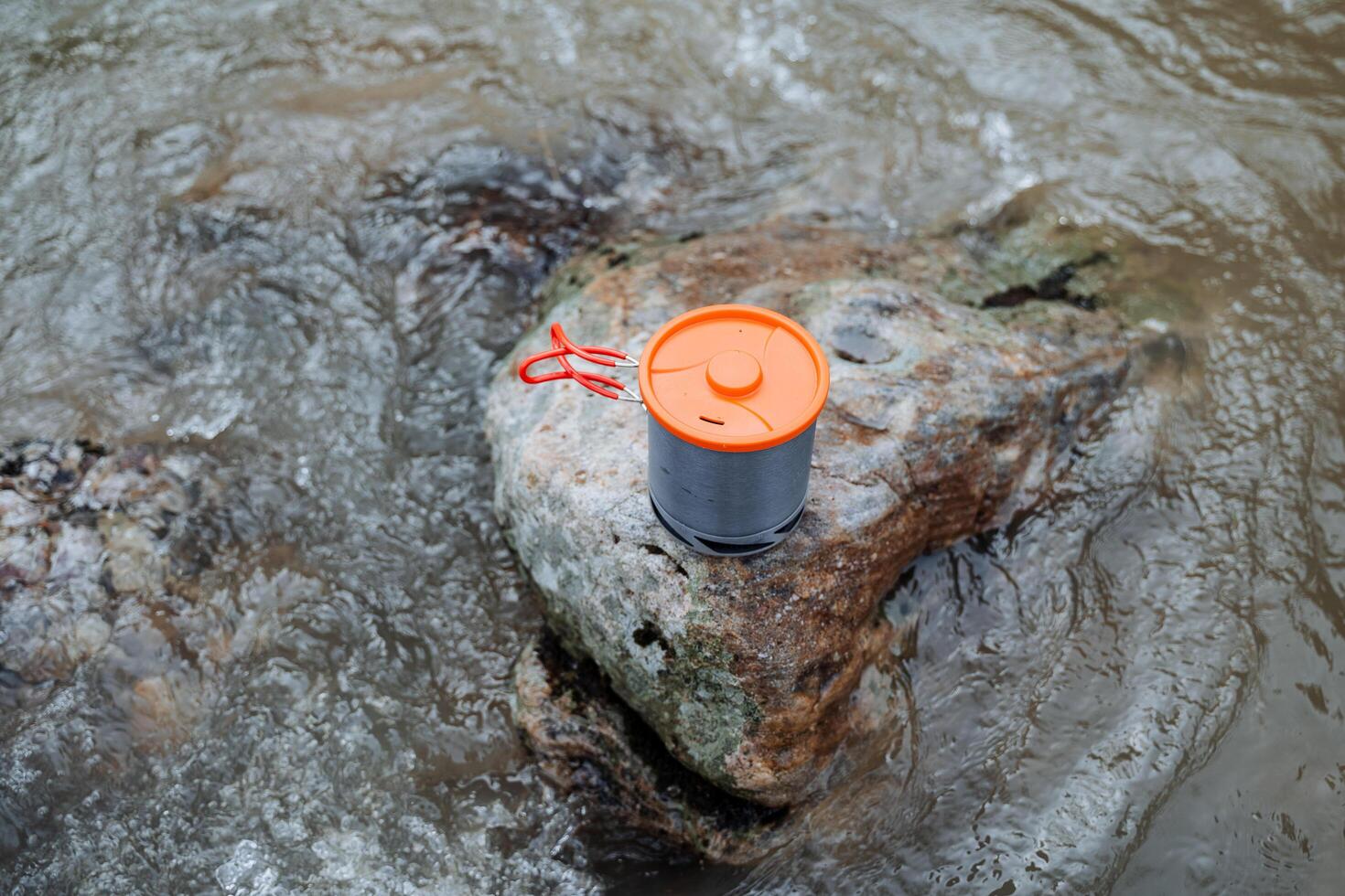 Tourist tableware concept on a stone against the background of water, a pot stands on a stone in the river, an orange cup lid, folding handles. photo