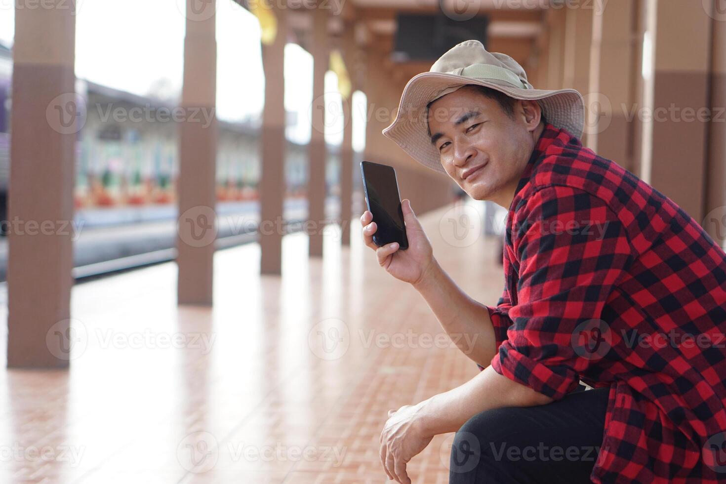 Asian man traveler is waiting someone at platform of train station, holds mobilephone to talk on the phone. Concept, travel alone, transportation, communication or connection photo