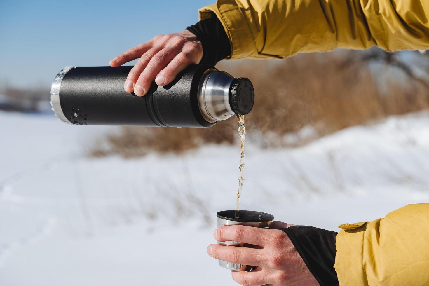 Pour the tea from the thermos into a mug. Water flows, pour a hot drink against the background of snow. Black metal thermos, flask for drinks. photo