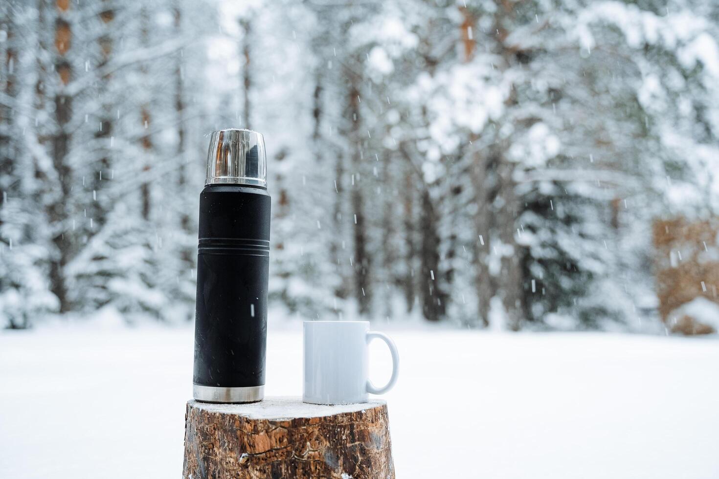 black matte thermos and white mug with hot drink on stump. Hiking and picnic staff. Drinking warm tea or coffee in the winter frost. Minimalistic subject shot photo