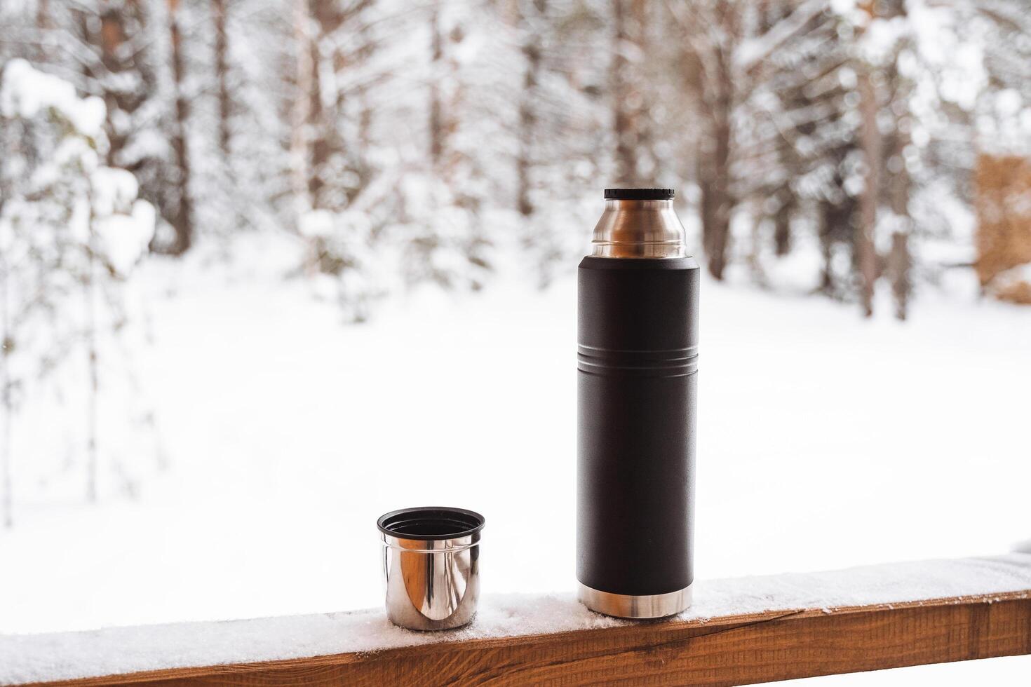 black matte thermos and mug with hot drink on window sill of the outdoor veranda. Hiking and picnic staff. Drinking warm tea or coffee in the winter frost. photo