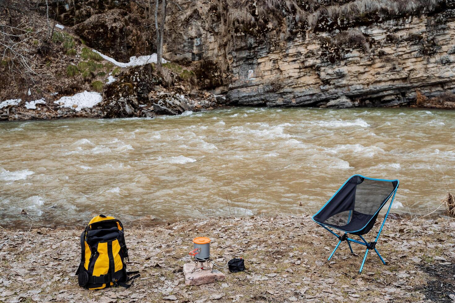 Rest on the bank of a mountain river, spring hike camping organization by the water, tourist equipment, backpack stands on the ground, cook food in a pot on a burner, folding chair photo