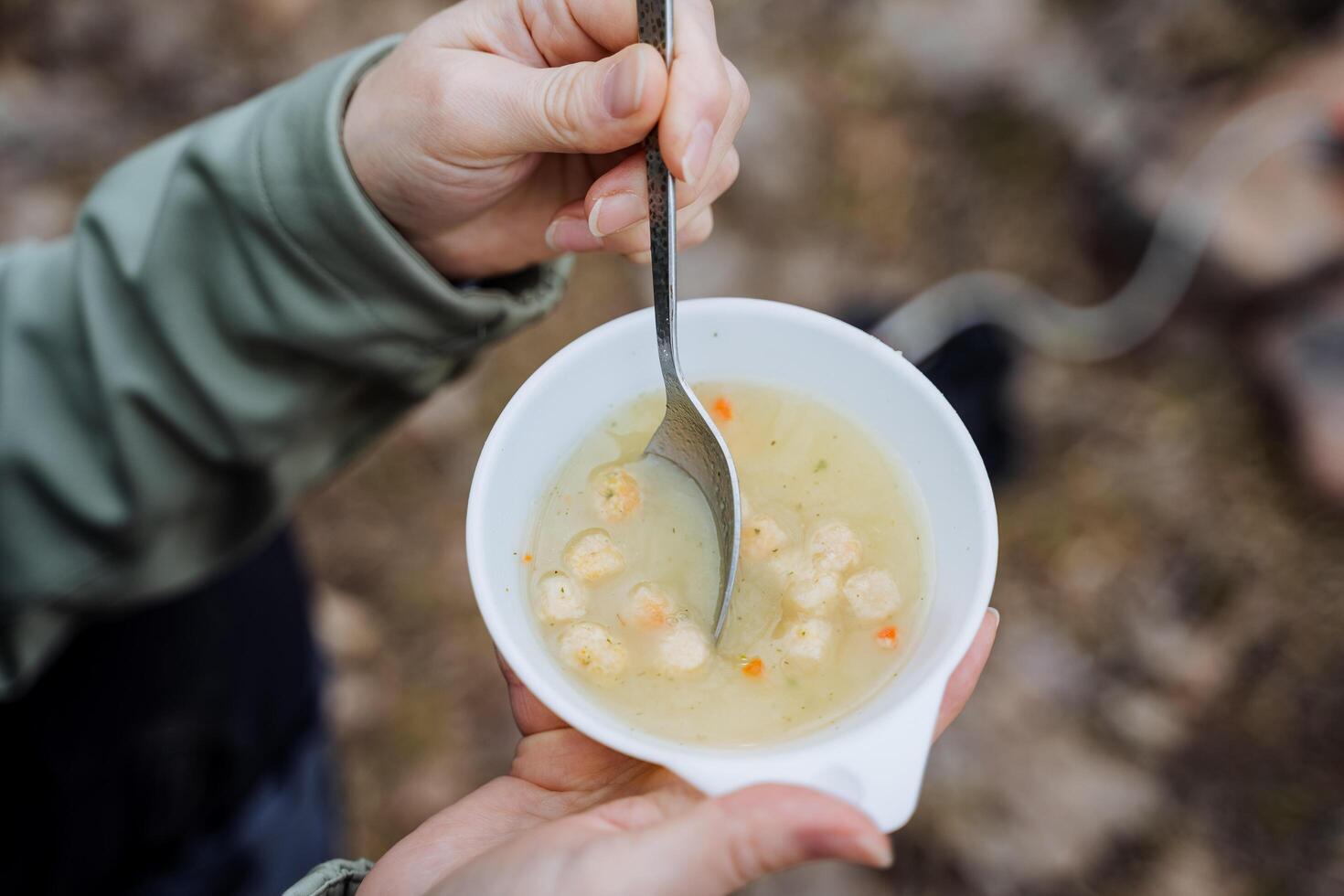 A bowl of soup is shot close-up, food on a hike, tourist breakfast in nature, potato soup is eaten with a spoon, hot food, a white plate, a girl's hands. photo