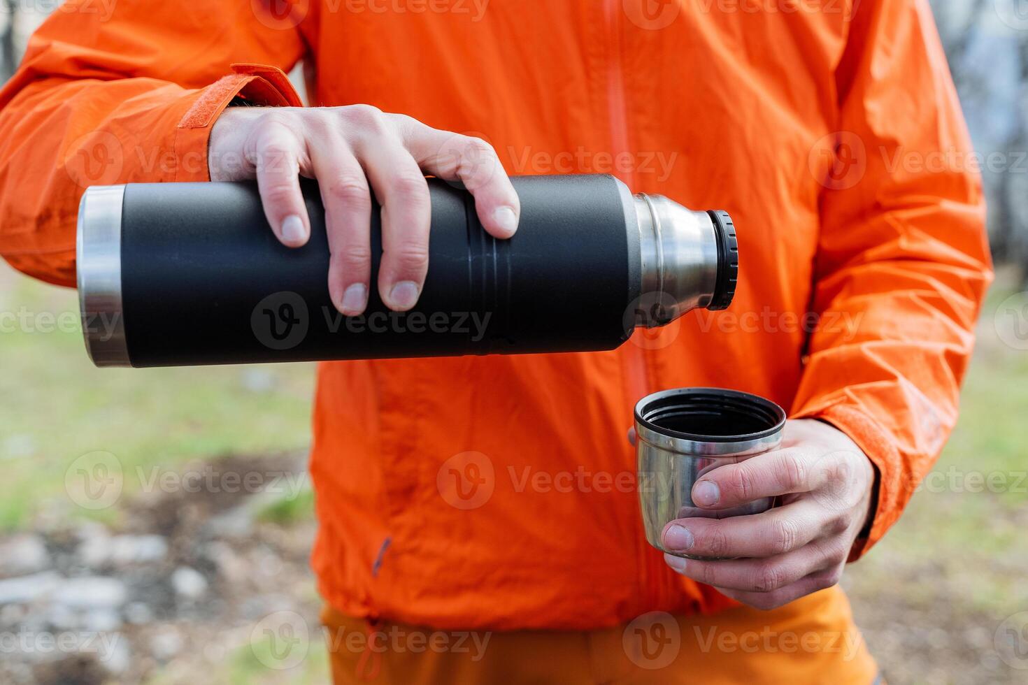 A tourist man seems to be holding a thermos with hot tea in his hands, a man pours coffee into a mug in nature, tourist equipment, hiking equipment, a vacuum bottle. photo