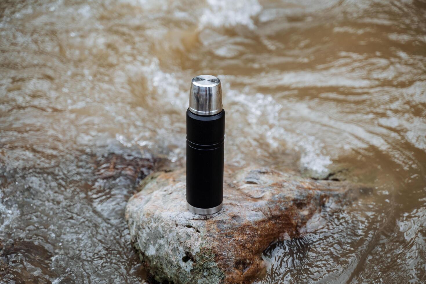 A vacuum thermos shot close-up stands on a rock in the river. The concept of a thermo coffee cup, trekking in the spring in the mountains. photo