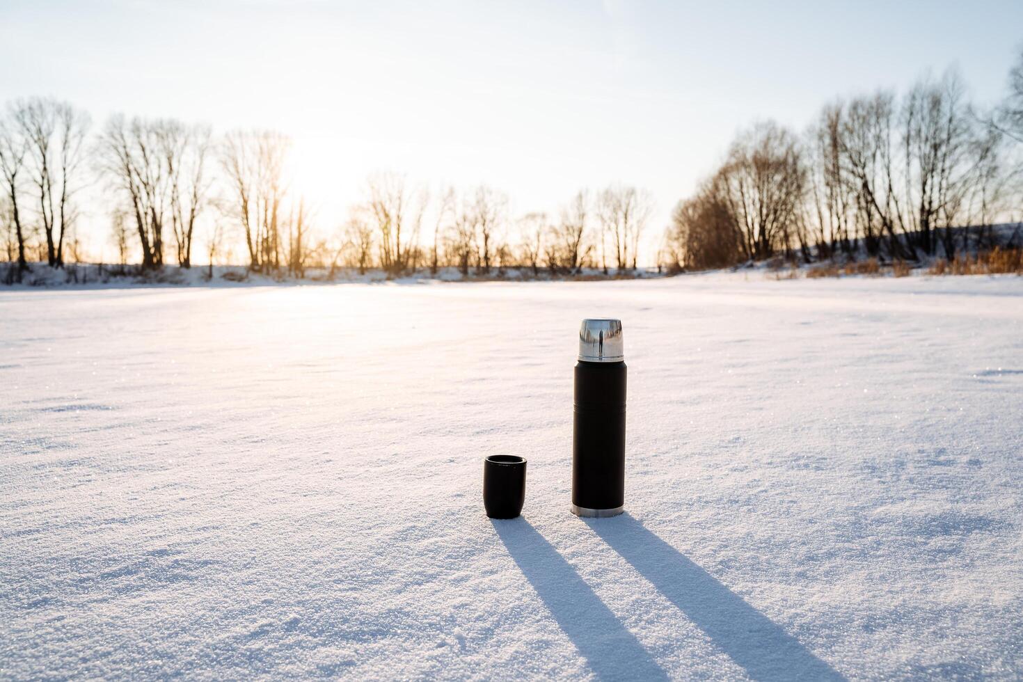 A thermos of black with a cup stands on the snow. Tourist utensils. Drink hot tea in winter to keep warm. Vacuum bottle for hot water. Glare of the sun in nature. photo