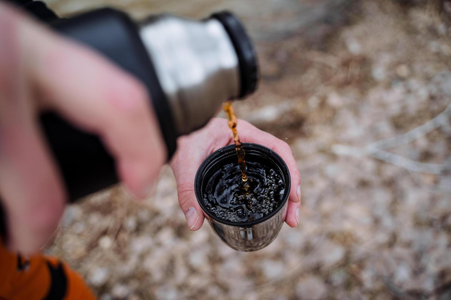 A thermos pour tea into a mug, a hand holds a glass of hot tea on a hike, bubbles in a cup, hot drinks in cold weather, tourist utensils, coffee in the morning. photo
