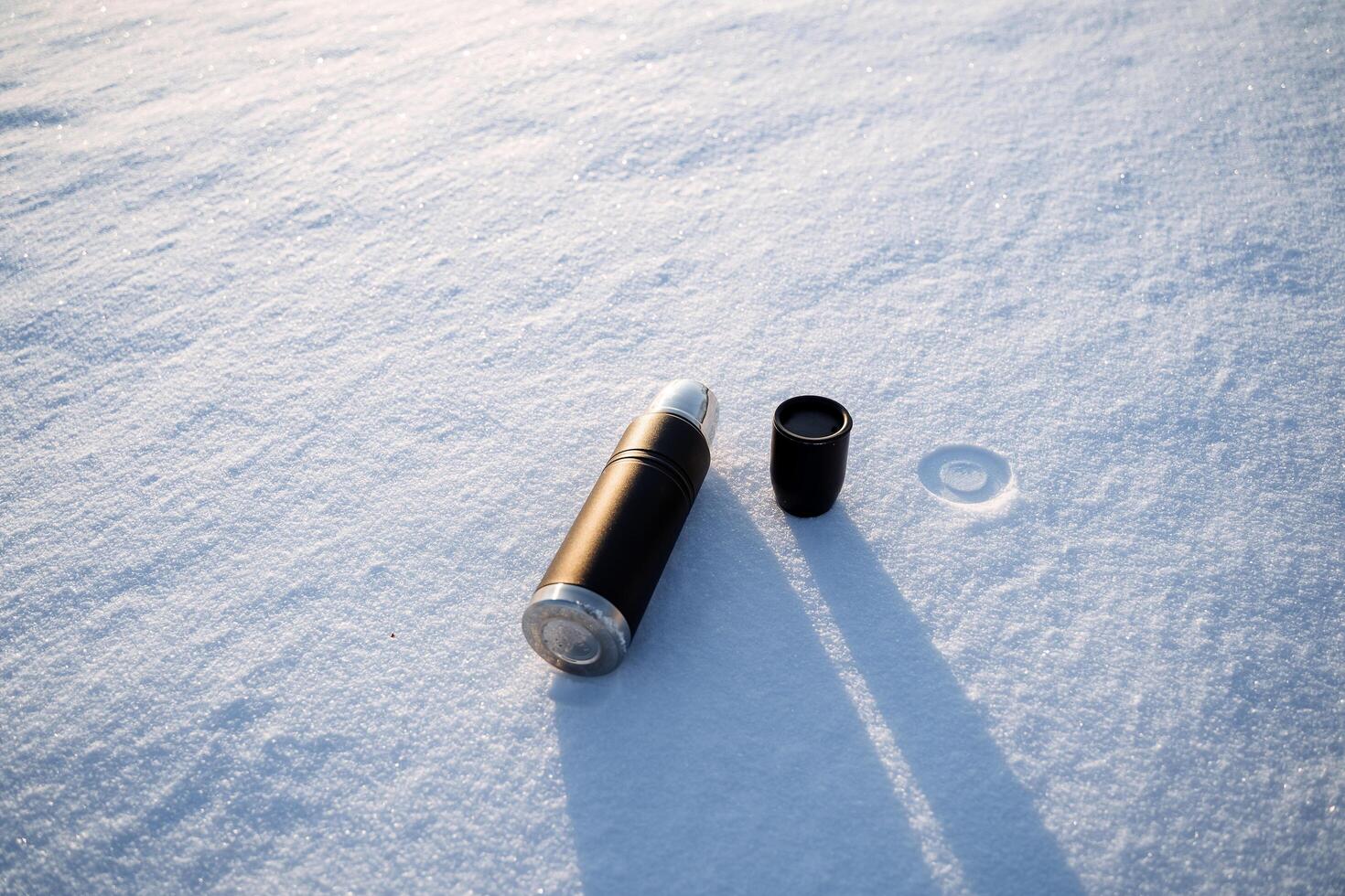 The thermos lies on the snow. The shadow of the dishes falls on the snow. A hot drink of coffee is poured into a thermal mug. photo