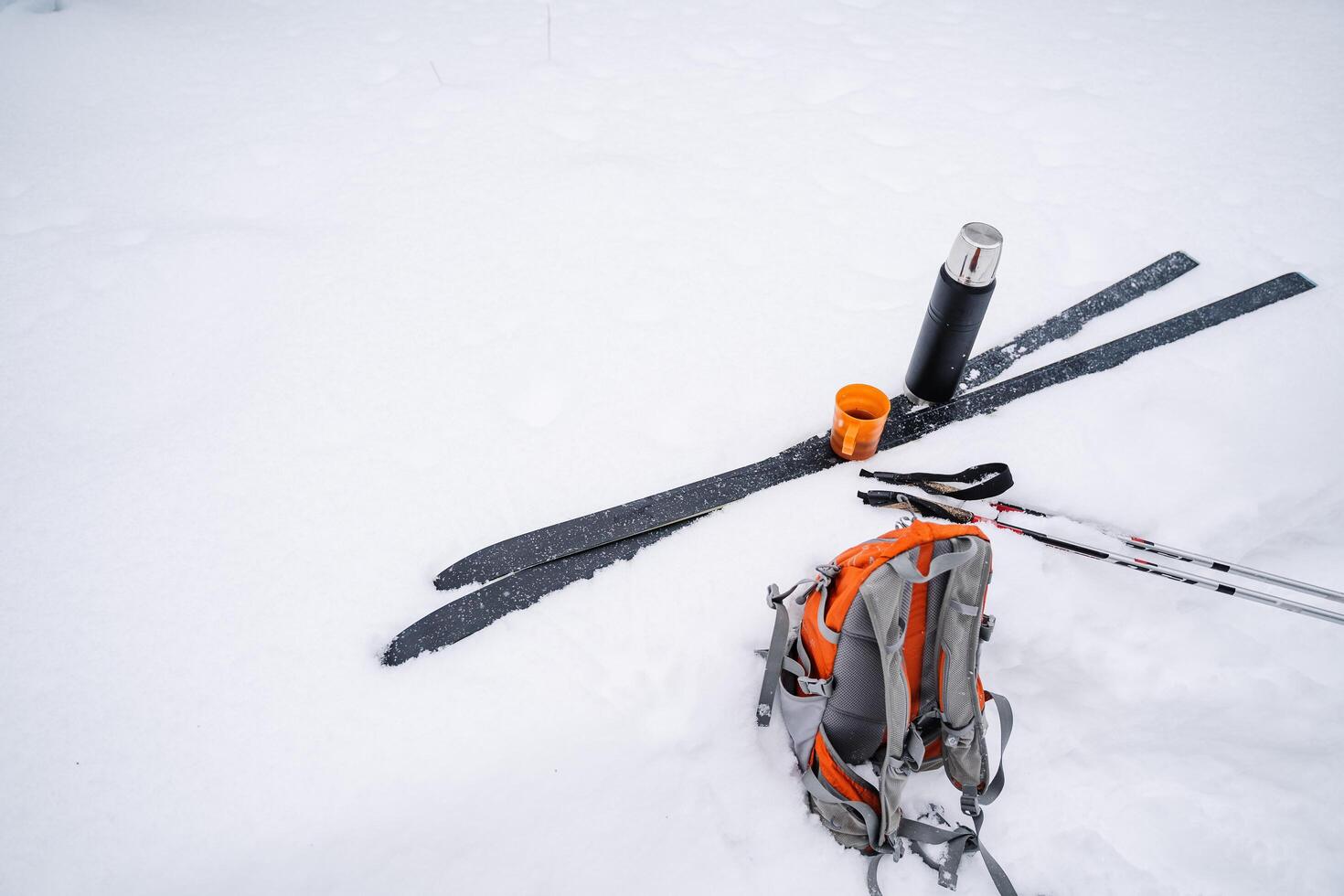 A black thermos with a mug of tea stand on skis in the snow. A bright orange hiking backpack is lying in the snow. Skiing in winter. photo