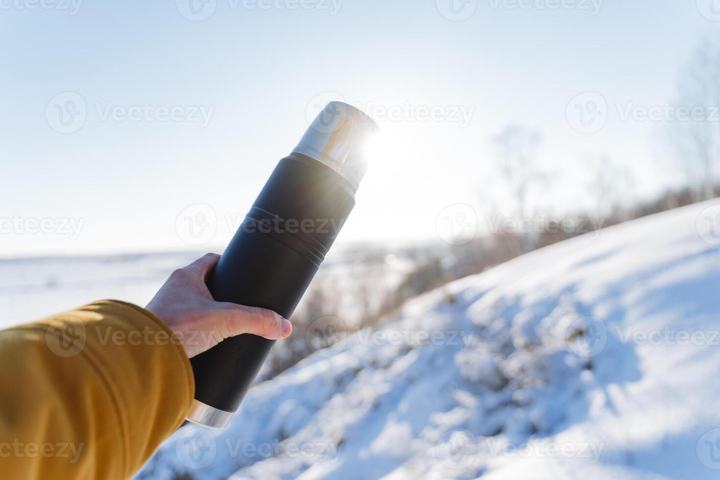 A hand holds a thermos of tea against the sky, sunlight glare into the camera. Black vacuum vessel bottle. The concept of winter trekking in the mountains. photo