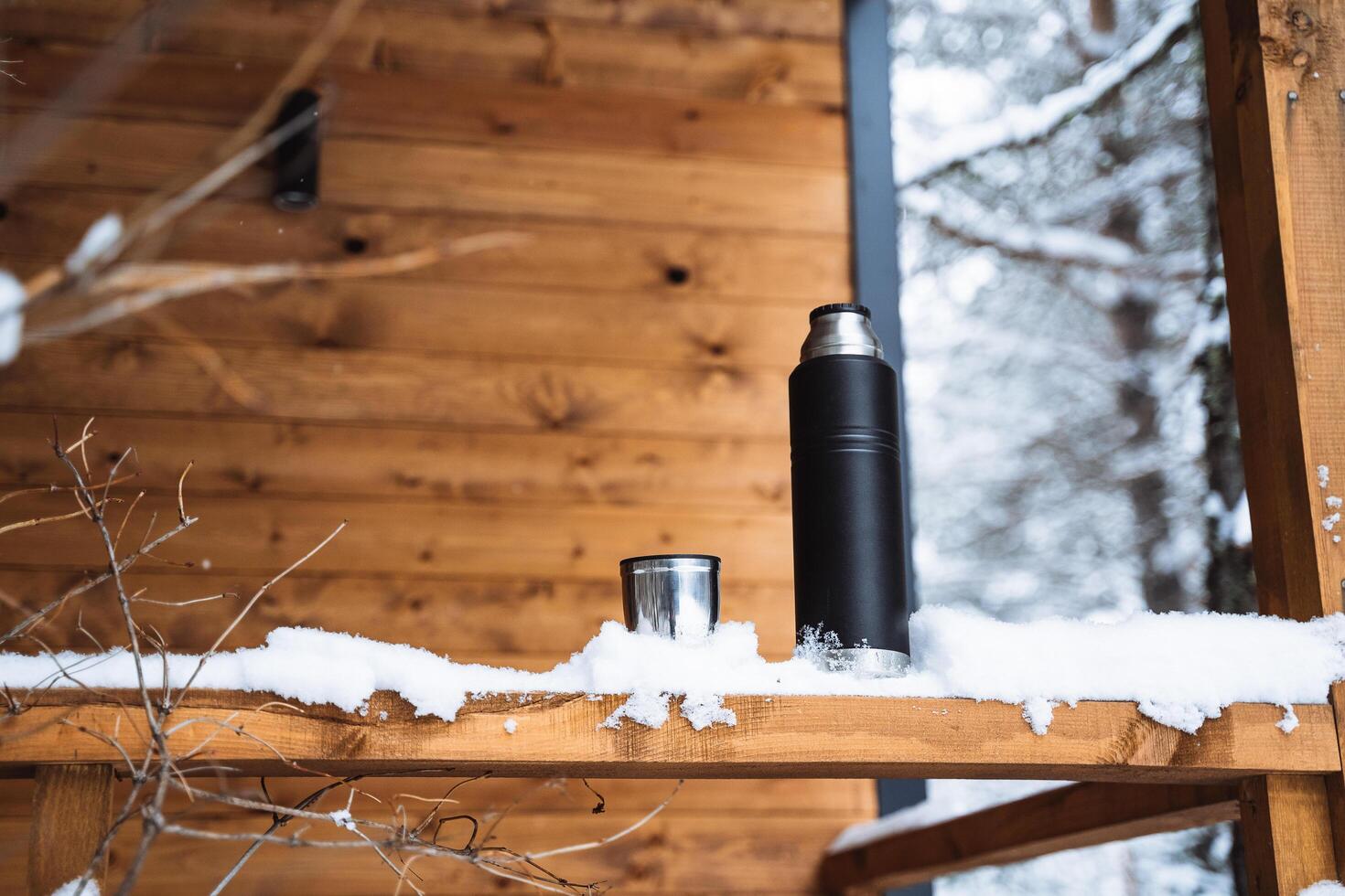 black matte thermos and mug with hot drink on window sill of the outdoor veranda. Hiking and picnic staff. Drinking warm tea or coffee in the winter frost. photo