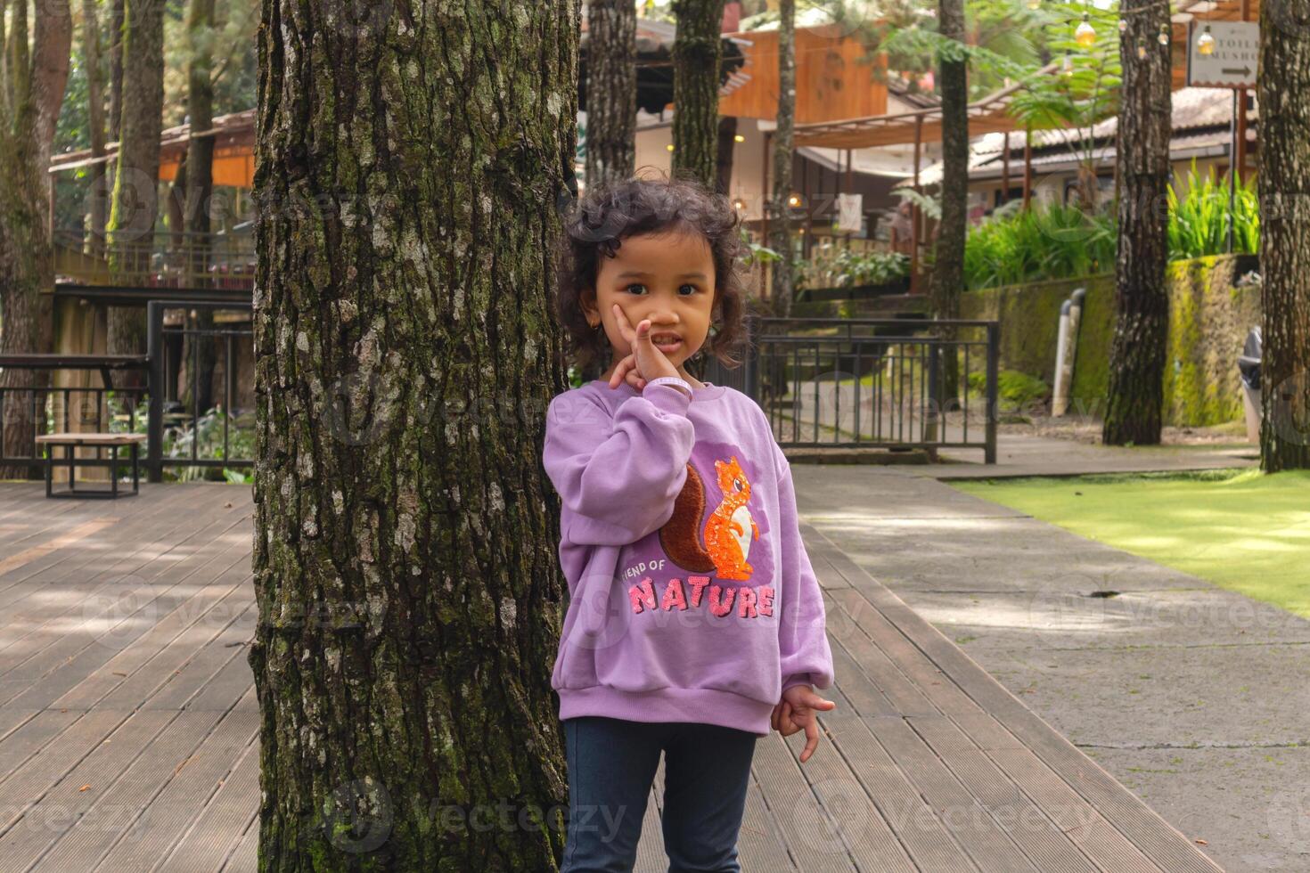 un asiático pequeño niña con Rizado pelo en un púrpura largo manga camisa en pie y posando con arboles o naturaleza antecedentes foto