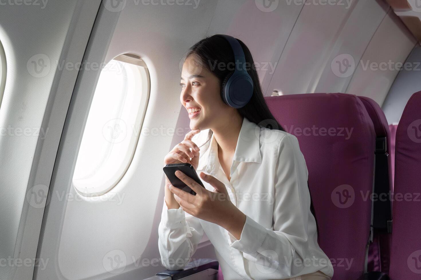 Cheerful Asian woman passenger in headphone watching online movie during intercontinental flight in cabin of aircraft, happy traveler using wifi connection on board photo