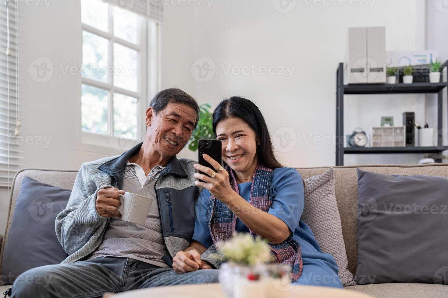 Happy elderly asian couple using smartphone sit on sofa doing ecommerce shopping online on website at home photo