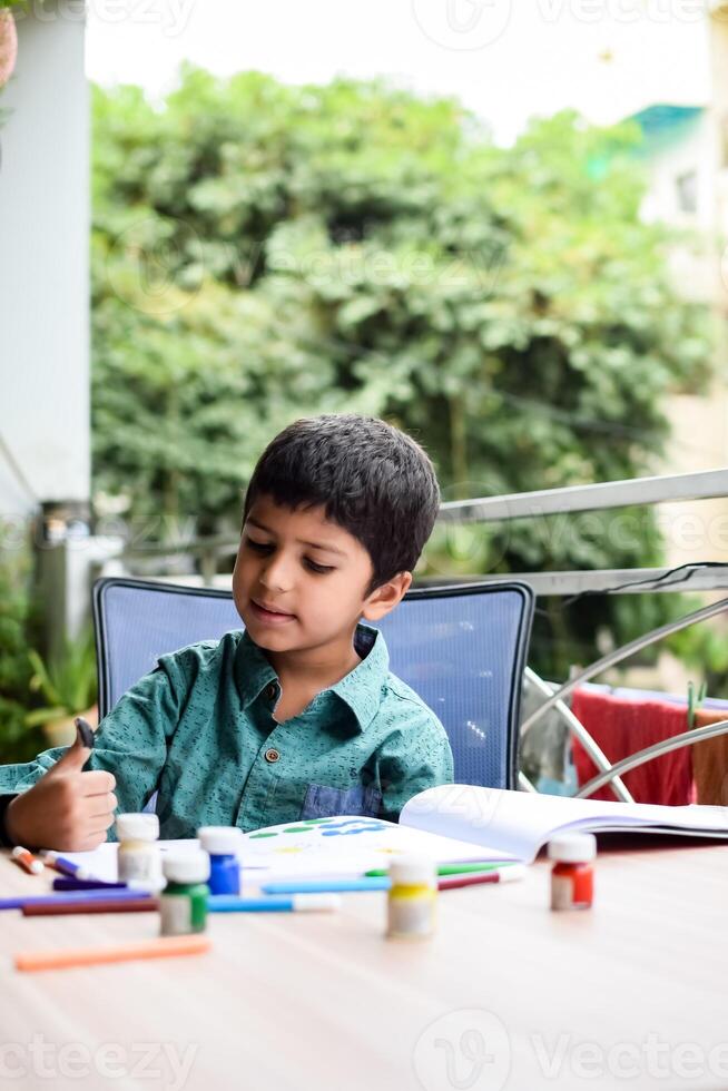 Smart Indian little boy perform thumb painting with different colourful water colour kit during the summer vacations, Cute Indian Kid doing colourful thumb painting drawing on wooden table photo