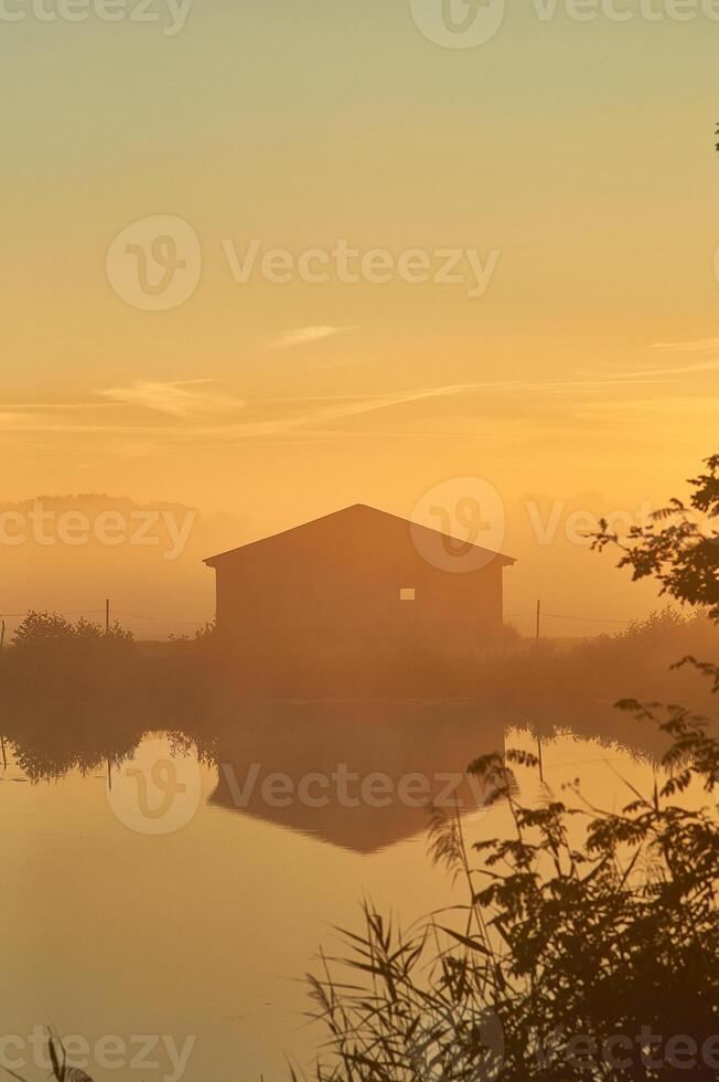 Old barn on lake shore on misty sunrise photo