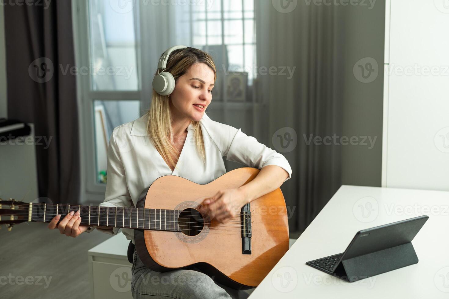 sereno joven mujer en inalámbrico auriculares utilizando acústico musical instrumento mientras sentado con ordenador portátil foto