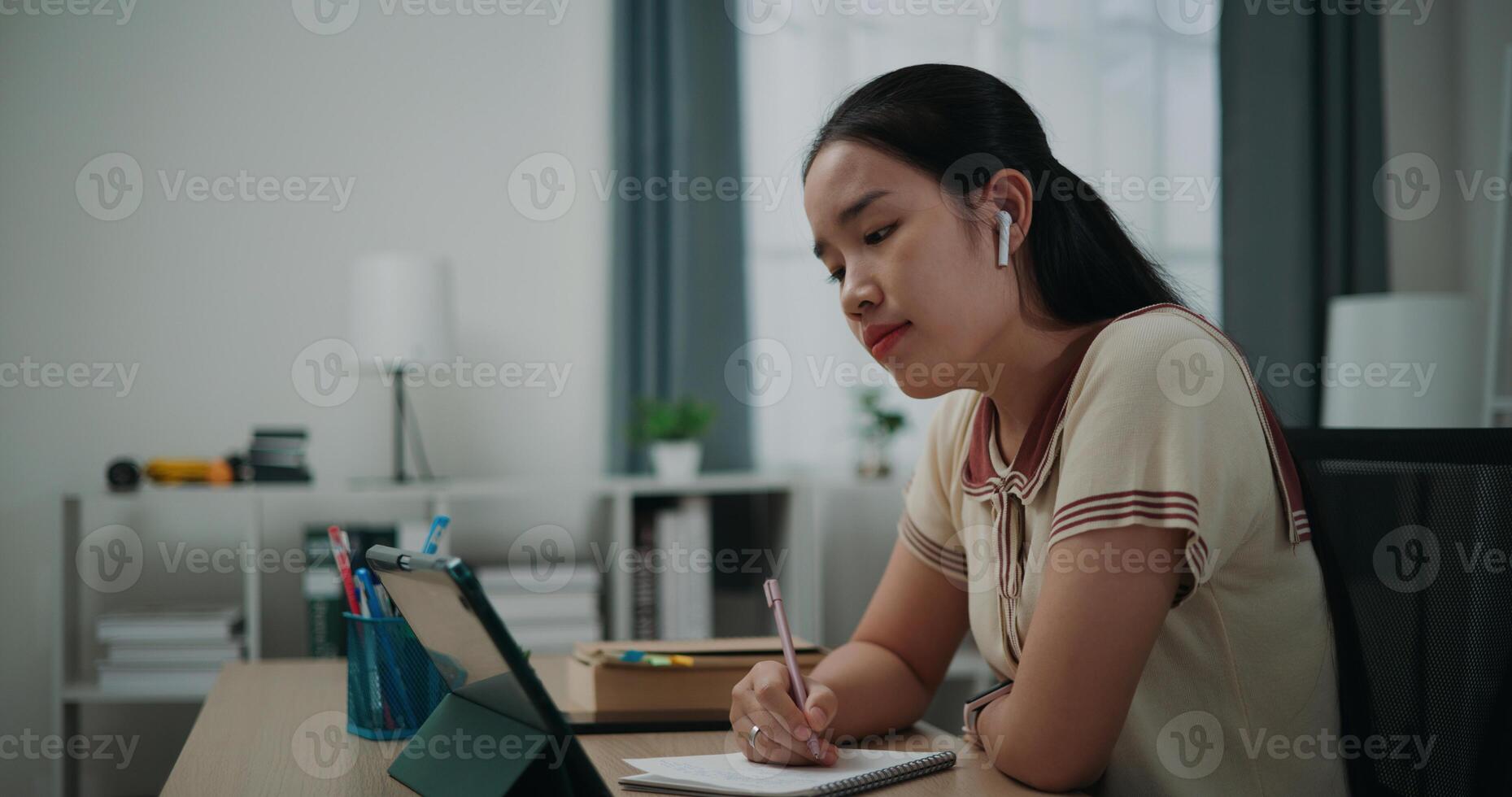 Selective focus, Side view, Female writer wear wireless headphones sitting at desk writing notes while working on tablet for online learning at home, creative thoughts to journaling photo