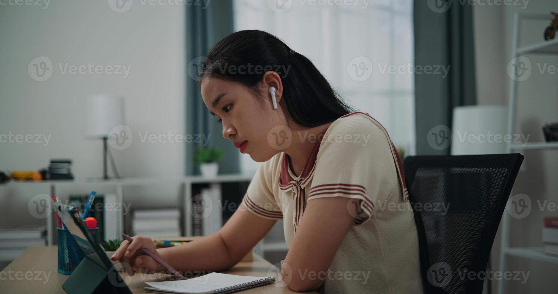Selective focus, Side view, Female writer wear wireless headphones sitting at desk writing notes while working on tablet for online learning at home, creative thoughts to journaling photo