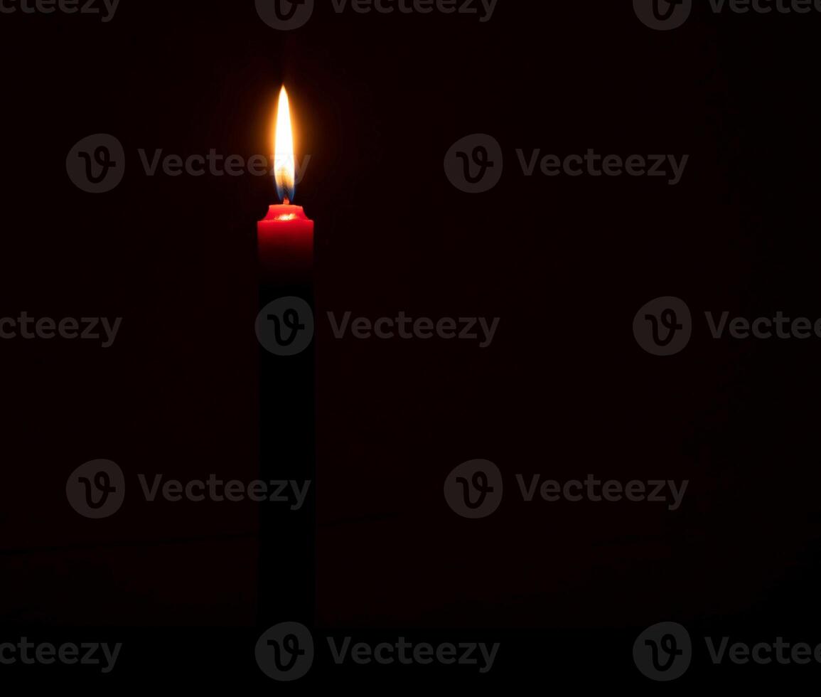Single burning candle flame or light glowing on a small red candle on black or dark background on table in church for Christmas, funeral or memorial service with copy space. photo