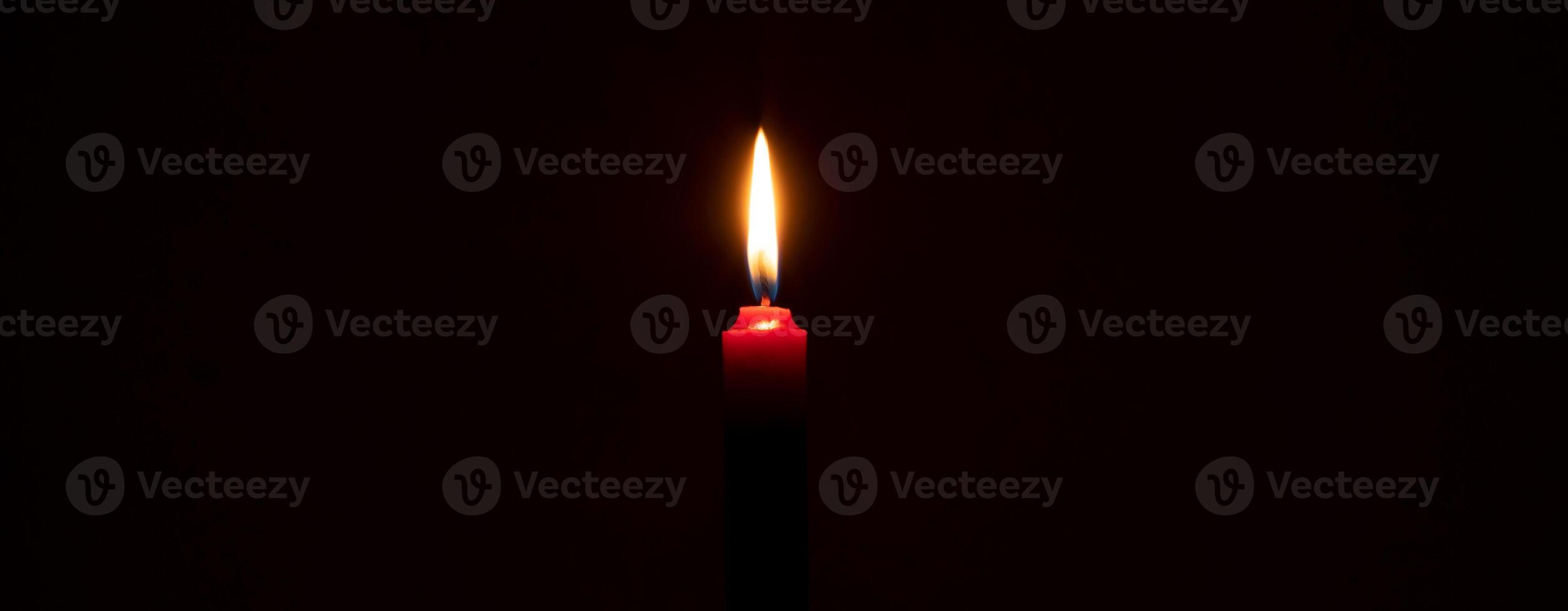 Single burning candle flame or light glowing on a small red candle on black or dark background on table in church for Christmas, funeral or memorial service with copy space photo