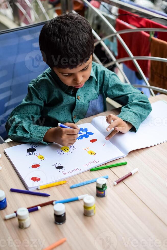 Smart Indian little boy perform thumb painting with different colourful water colour kit during the summer vacations, Cute Indian Kid doing colourful thumb painting drawing on wooden table photo