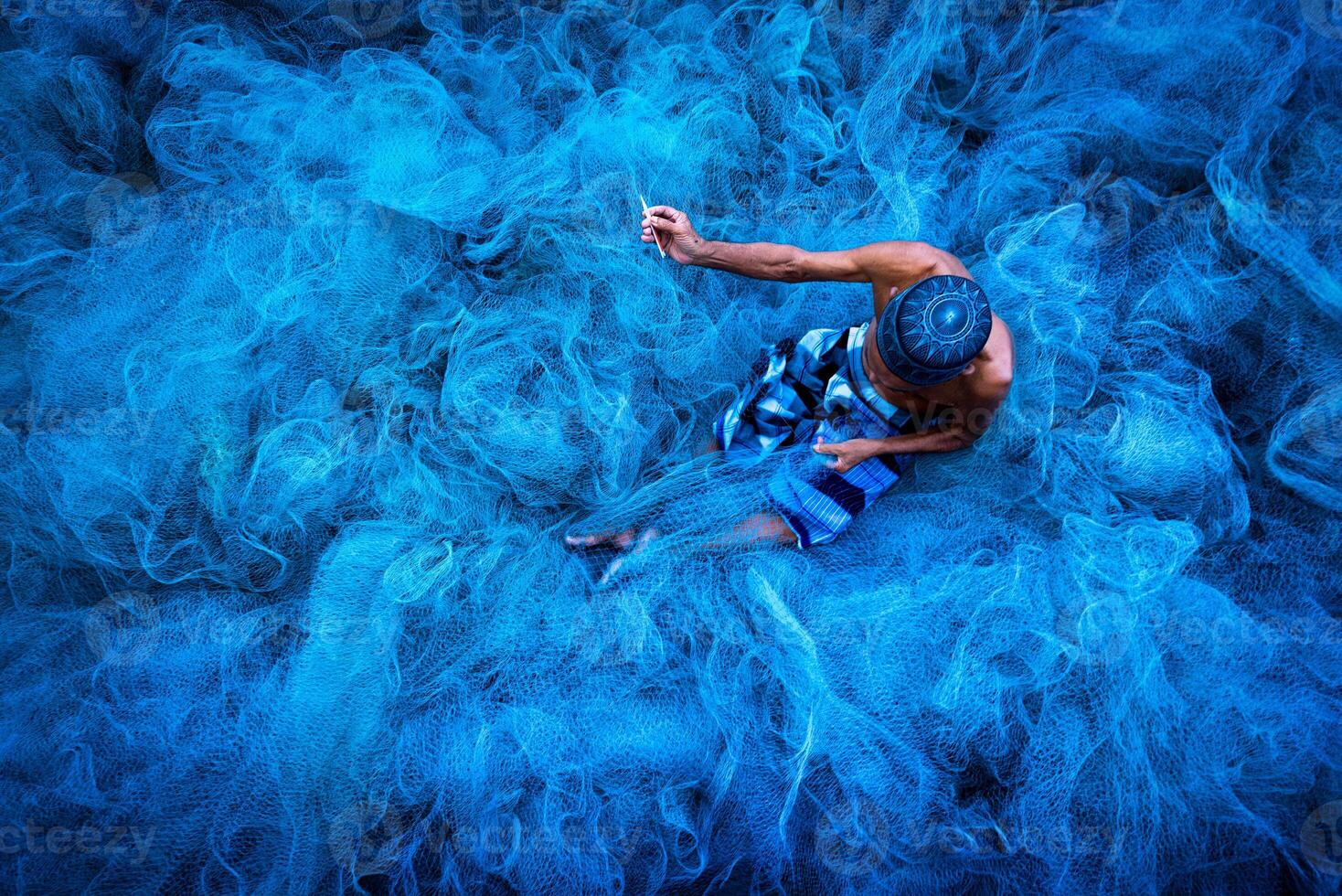 Old fisherman hands sewing blue fishing nets sitting on the ground and surrounded big net photo