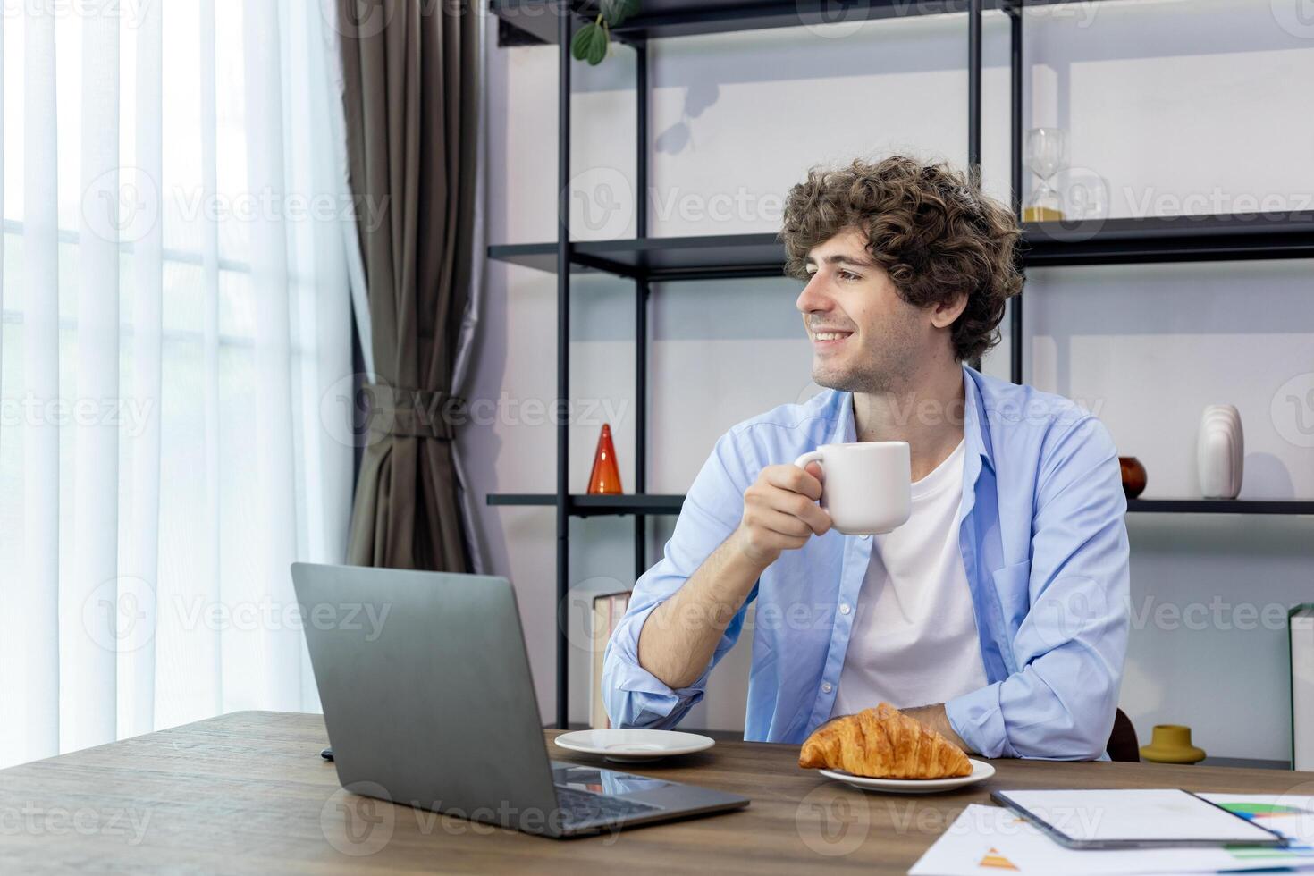 caucásico hombre es trabajando a su hogar oficina mientras Bebiendo café y comiendo cuerno para desayuno para Lanza libre y empresario negocio foto