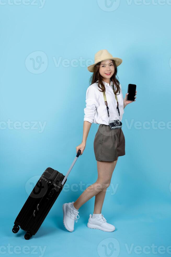 Beautiful young Asian tourist woman walking with trolley suitcase and smartphone, studio shot blue color isolated background photo