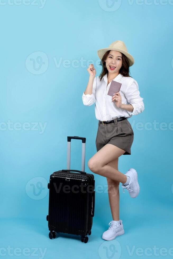 Excited Asian female traveler wearing summer clothes with suitcase, passport, do winner gesture isolated on blue background. photo