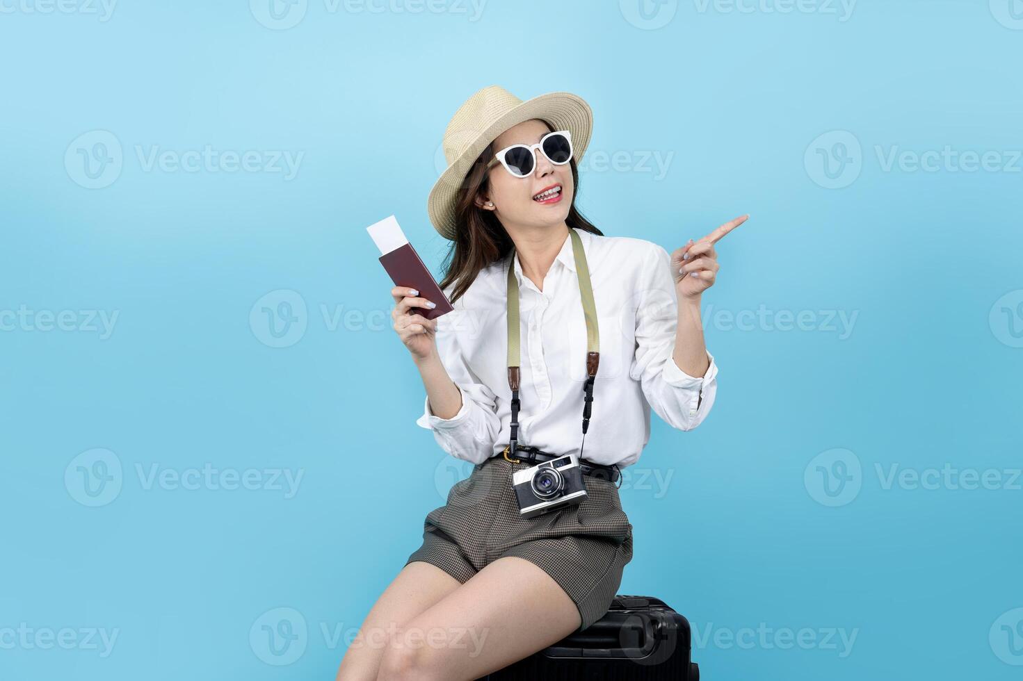 Happy smiling young Asian tourist woman sitting on luggage holding passport and hand pointing to copy space going to travel on holidays photo