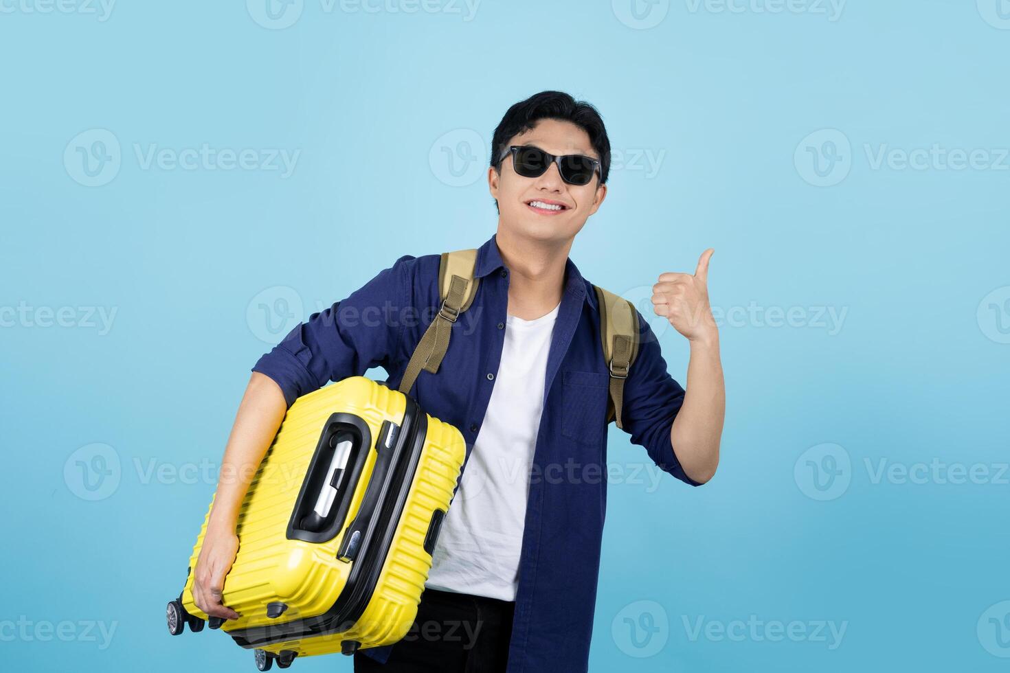 Excited handsome Asian male tourist in casual clothes hat backpack holding suitcase bag, shows thumb up gesture on blue background. photo