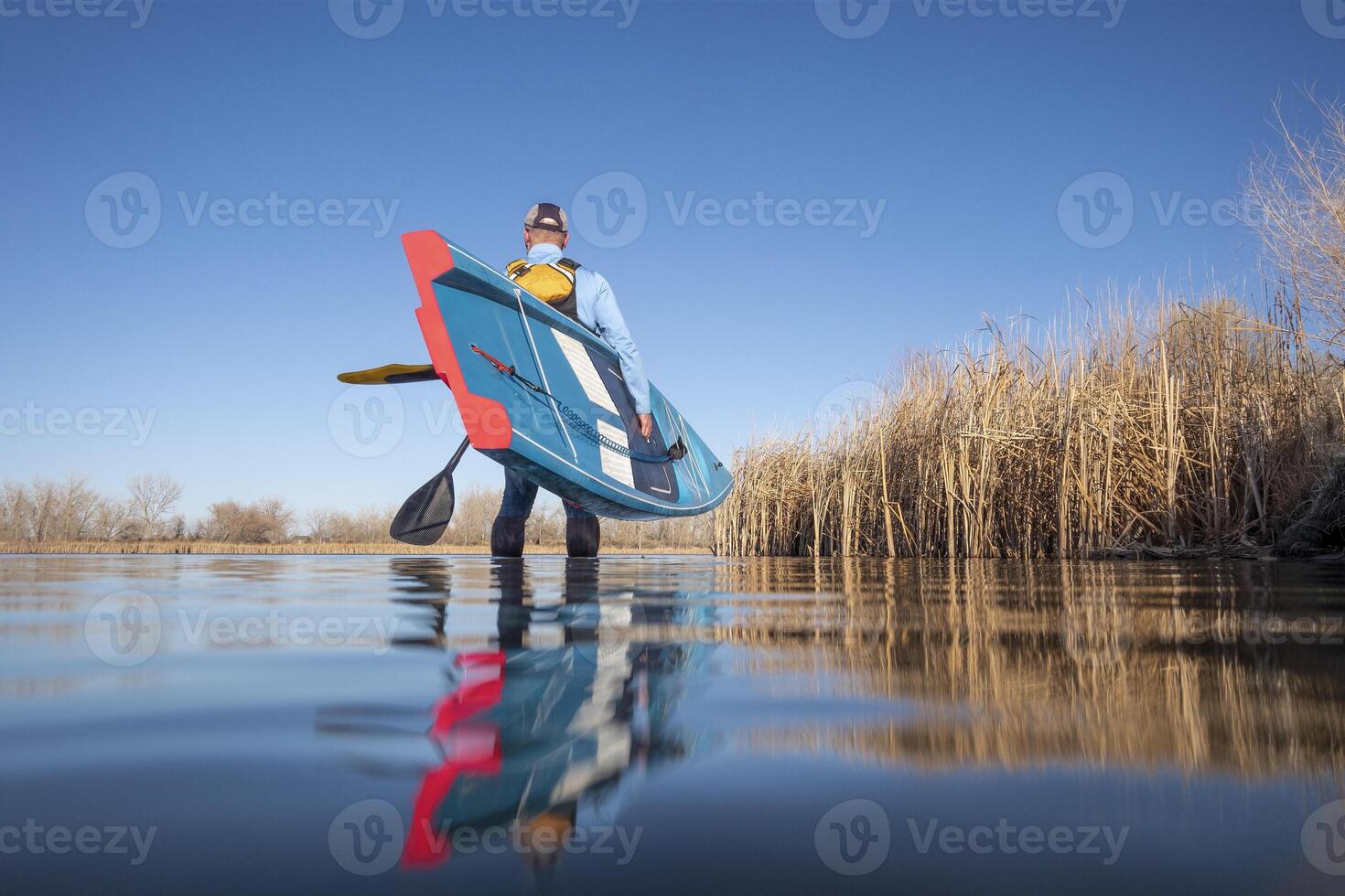 senior male paddler is launching a stand up paddleboard on a calm lake in early spring, frog perspective from an action camera at water level photo