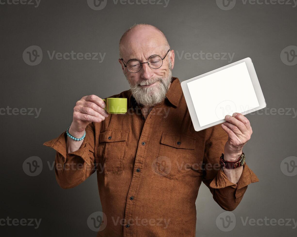 sonriente mayor hombre es Bebiendo café y demostración un blanco digital tableta foto