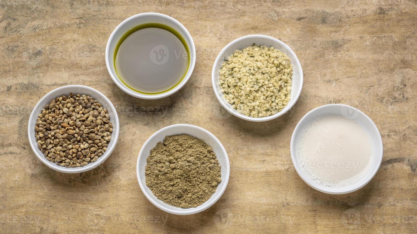 collection of hemp seed products, hearts, protein powder, milk and oil in small white bowls against textured bark paper, superfood concept photo