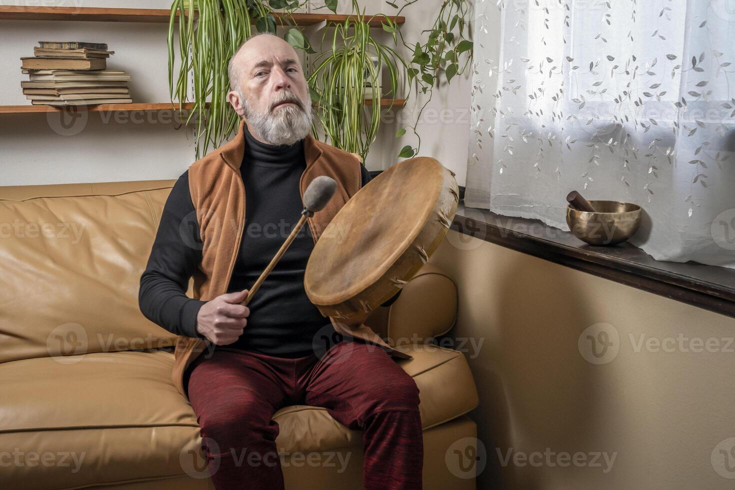 senior man is playing a native American style, shaman drum with a beater at home, lifestyle and hobby concept photo