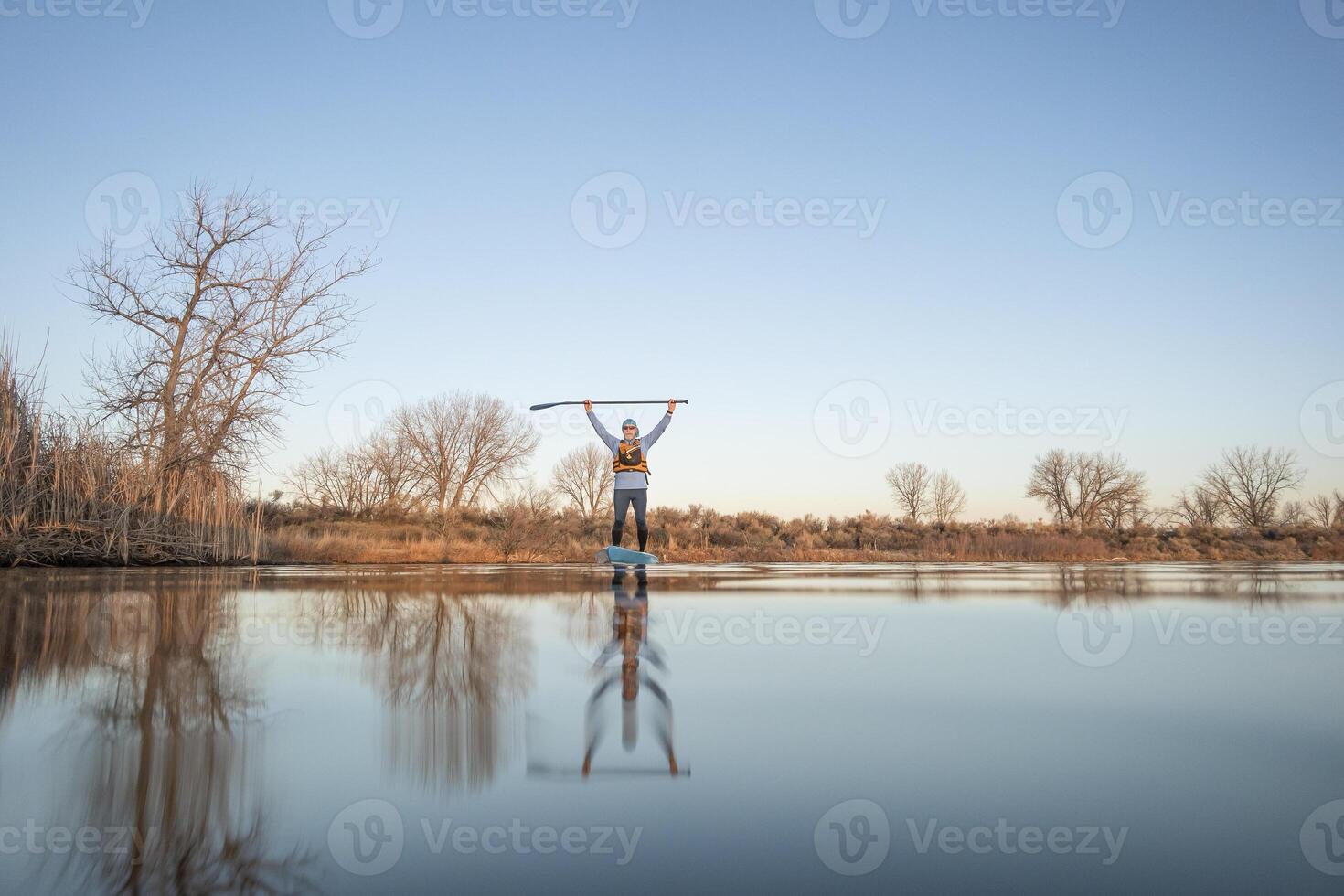 contento mayor palista es remar estar arriba paddleboard en calma a puesta de sol en temprano primavera en fuerte collins, Colorado lago, rana perspectiva, parcialmente sumergido acción cámara foto