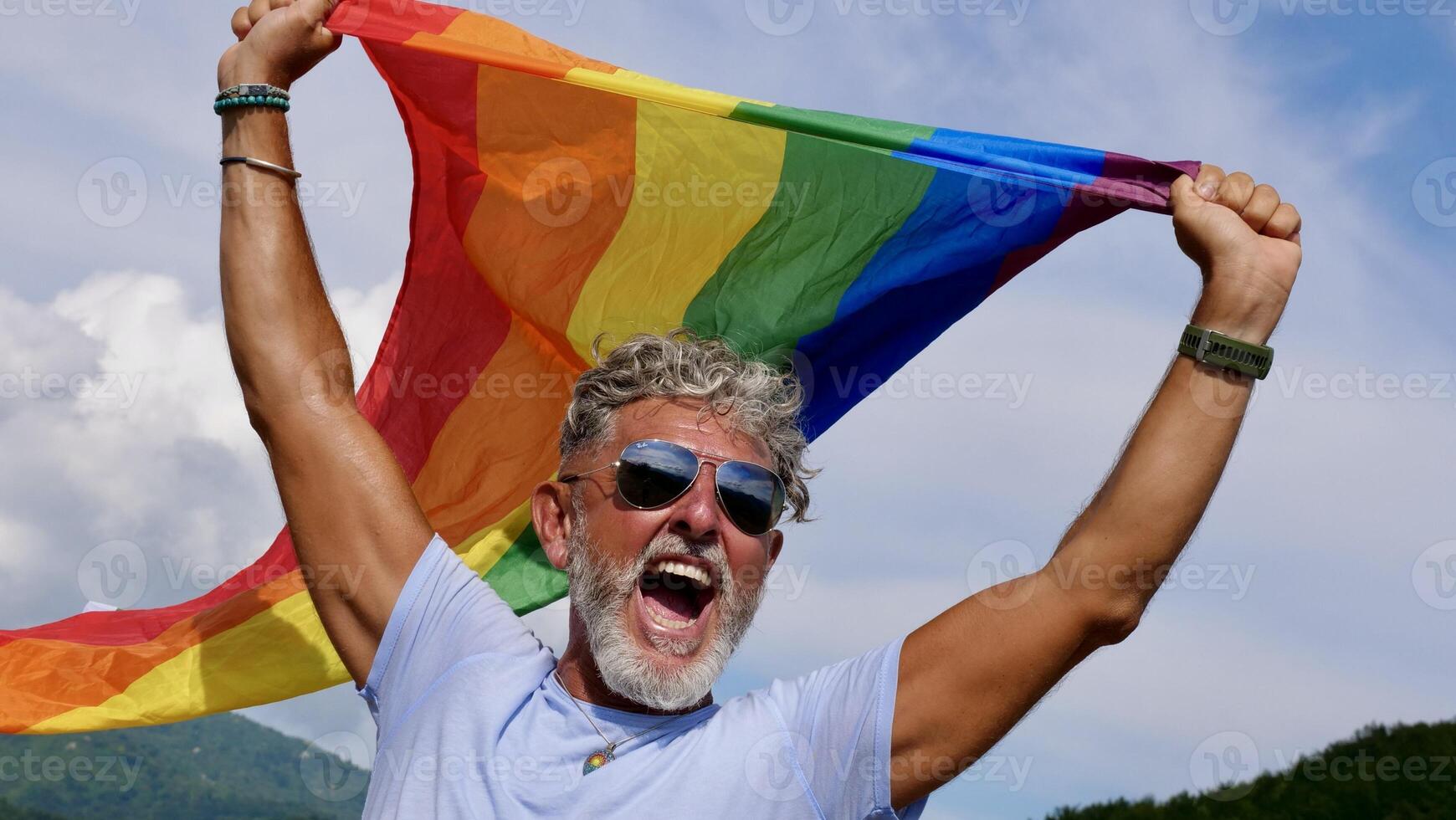 Portrait of a gray-haired elderly Caucasian man with a beard and sunglasses holding a rainbow LGBTQIA flag against a sky background, shouts in protest, Celebrates Pride Month Coming Out Day photo