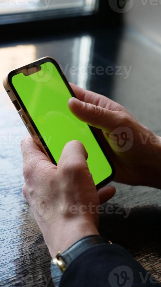 Close-up smartphone with green screen in men's hands. A businessman scrolls the phone with chroma key in the office. Vertical photo