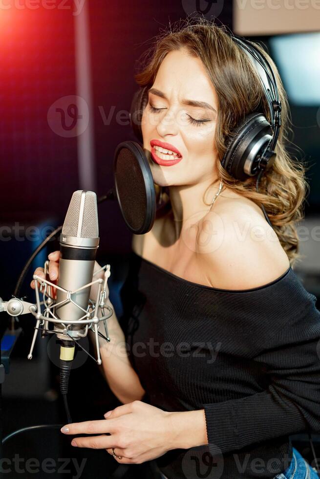 Young woman with the professional microphone in the recording studio. Vocalist girl in the studio. Beautiful singer in headphones sings into microphone with closed eyes. Closeup. photo